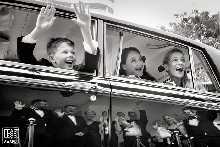 Children smiling and waving from a car window during a wedding celebration with guests reflected on the car door in a memorable wedding photo.