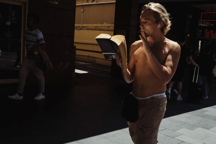 Shirtless young man reading a book and smoking on a sunlit street in candid street photos by Alex McClintock.