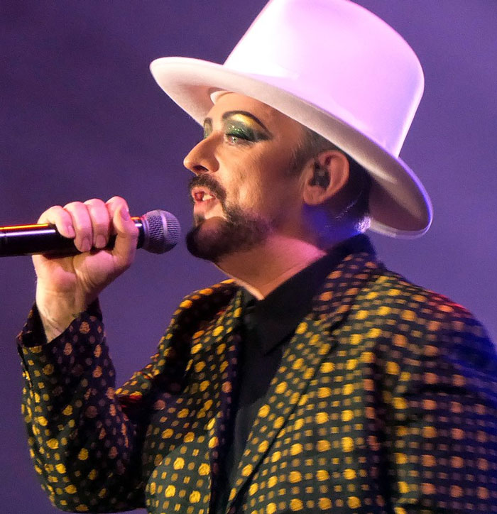 Male performer with glittery eye makeup and white hat singing into a microphone, representing famous people who did evil things.