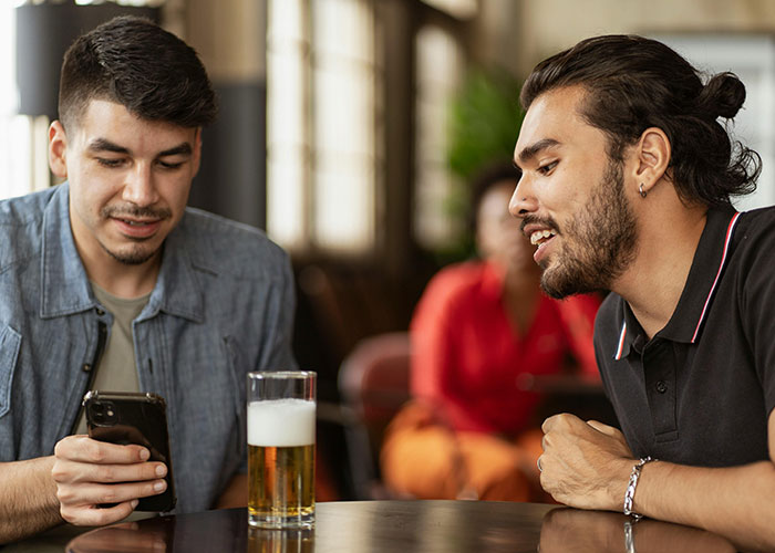 Two men at a bar during an awkward date, one looking at his phone while the other talks, with a beer on the table.