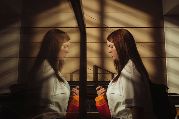 Young woman with red hair holding an orange bottle, reflected in glass, captured in candid street photos by Alex McClintock.