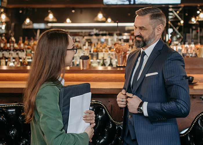 A man in a suit and a woman with glasses having an awkward date conversation at a bar with bottles behind.