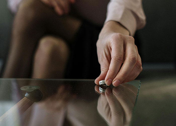 Person sitting on a couch, holding an engagement ring near a glass table, symbolizing called off engagements.