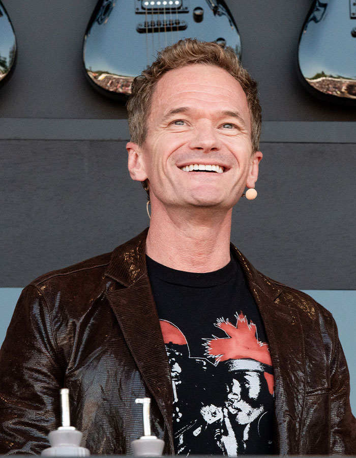 Smiling male celebrity wearing a dark jacket and graphic t-shirt, appearing youthful and vibrant on stage with guitars behind.