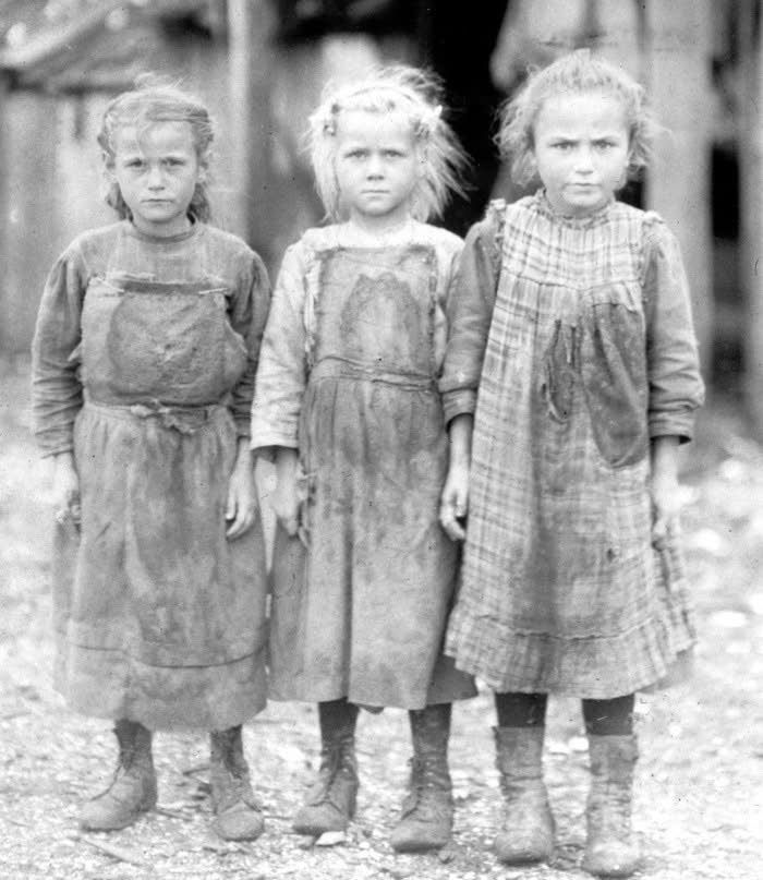 Three young girls in worn, dirty dresses standing outdoors in a vintage black-and-white historical photo.