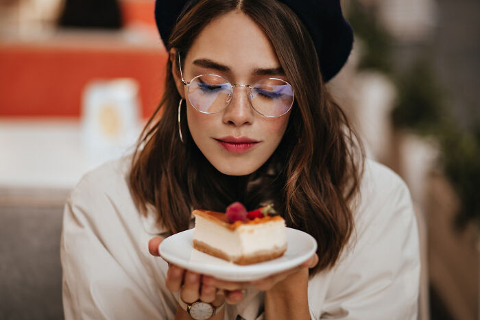 Young woman wearing glasses and a beret, closely examining a cheesecake slice, showing commitment to a lie concept.