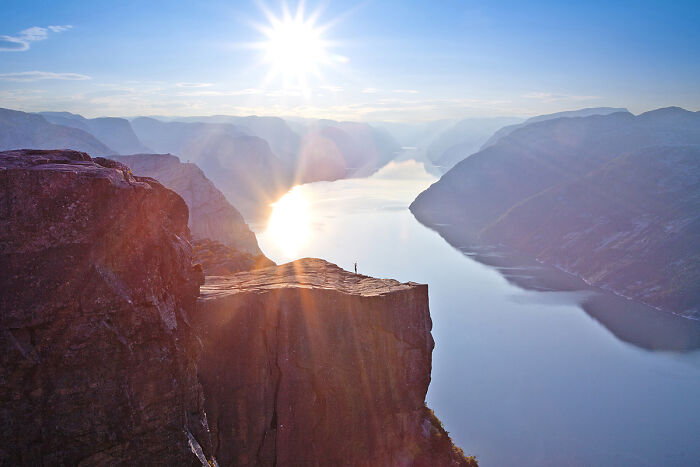 Person standing on cliff edge overlooking a fjord at sunrise in breathtaking travel photos captured by the community