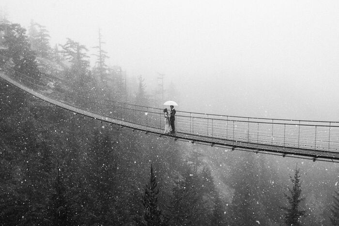 Couple sharing a tender moment under an umbrella on a snowy suspension bridge in a stunning wedding photo.