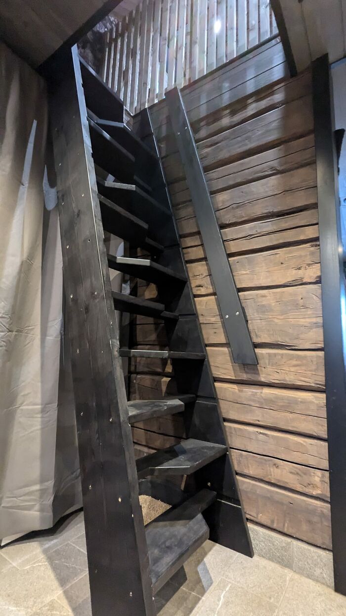 Steep narrow black wooden death stairs leading up inside a rustic wooden cabin with bright natural light.