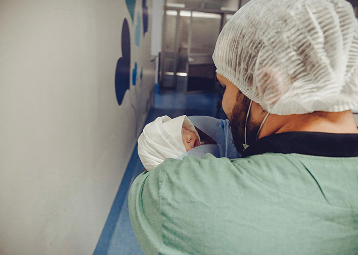 A dad wearing scrubs and a hairnet holding a newborn baby inside the birthing room after delivery.
