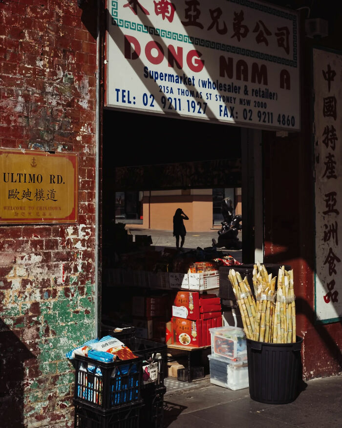 Candid street photo of a Chinatown supermarket exterior with vibrant produce and shadowed figure in the background.