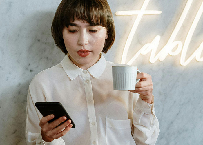 Young woman in a white shirt holding a cup and looking at her phone during an awkward date at a café.