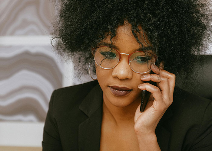 Woman with glasses and curly hair in a black blazer speaking on phone, reflecting on engagement and relationship challenges.