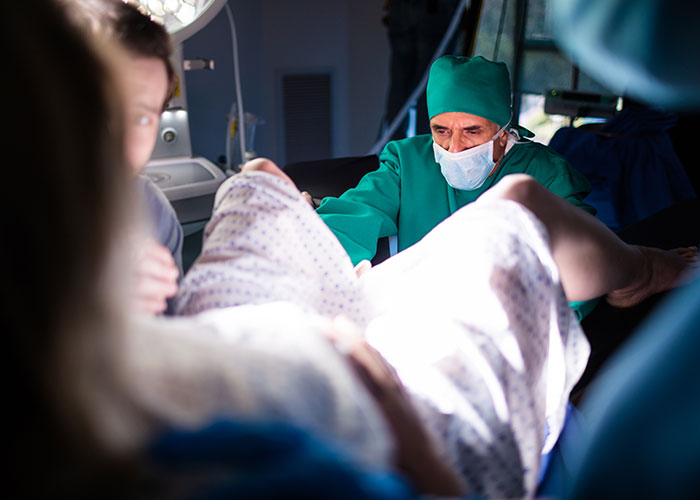 Doctor wearing surgical scrubs assisting a mother during childbirth with a dad supporting her in the birthing room.