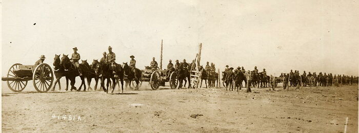 Long line of soldiers with horse-drawn artillery in a barren desert landscape, representing historical events.