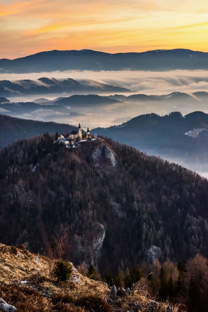 Scenic travel photo of a church atop a forested mountain with misty hills and colorful sunrise sky in the distance.