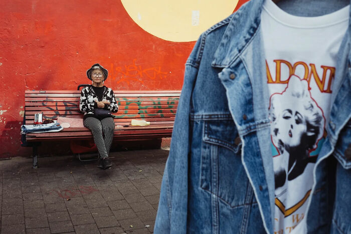 Elderly woman sitting on a bench against a red wall, captured in a candid street photo by Alex McClintock.