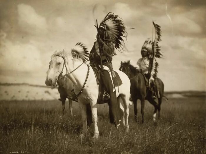 Native American warriors in traditional headdresses riding horses in an open field in a historical photo.