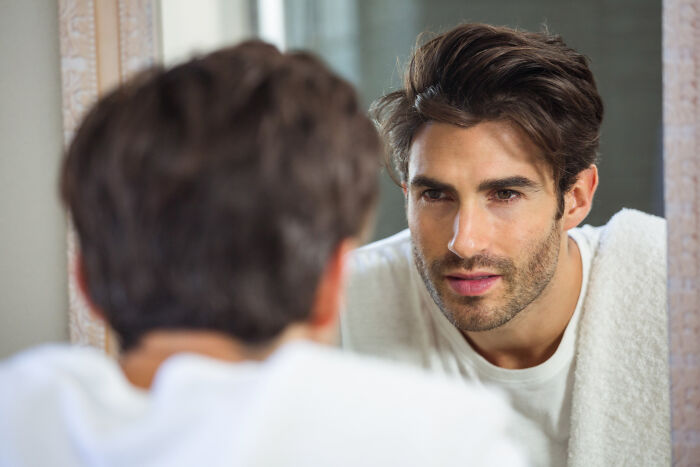 Man with dark hair and a white shirt looking intently at his reflection in the mirror, portraying a completely legal total psychopath.