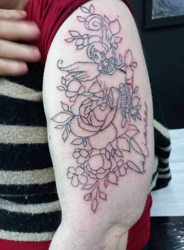 Outline tattoo of a fairy with flowers on upper arm showing a hilariously bad tattoo in need of improvement.
