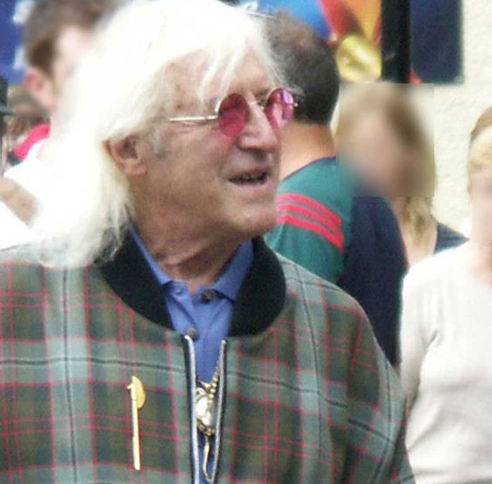Older man with white hair and pink glasses smiling in a crowd, representing famous people who did evil things in the past.