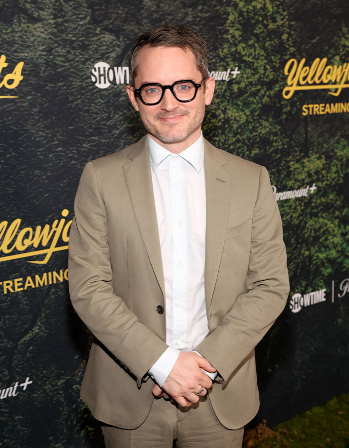 Man in beige suit and glasses posing at an event, one of the celebrities who look way younger than they actually are