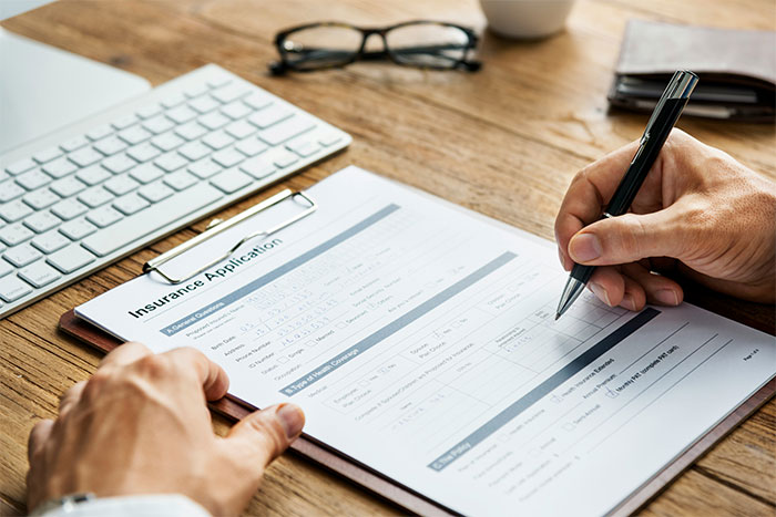 Person filling out an insurance application form on a clipboard at a desk with keyboard and glasses nearby. Person filling out an insurance application form on a clipboard at a desk with keyboard and glasses nearby.