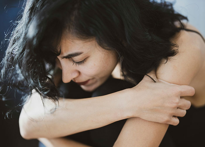 Woman with black curly hair looking down, embracing herself, expressing sadness and emotional distress indoors.