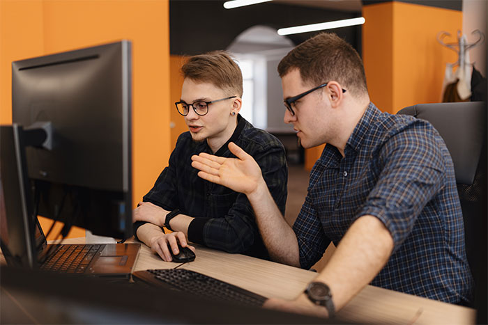 Two employees discussing deployment rule details intently while working at computers in a modern office setting.