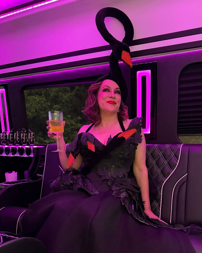 Jennifer Tilly wearing a black swan-themed outfit, holding a drink, seated in a purple-lit luxury limousine interior.