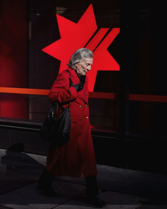 Elderly woman in a red coat walking past a bright red star, a candid street photo capturing everyday urban life.