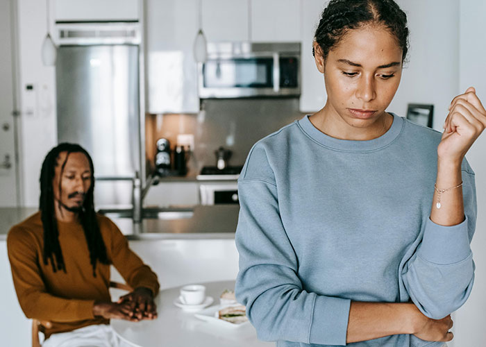 Young woman looks upset and distant while man sits behind her in a kitchen, reflecting on engagement called off over cheating with best man.