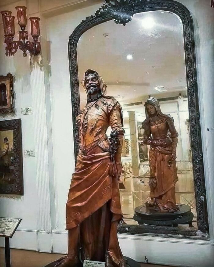 Statue of a man in historical attire with a woman’s reflection in a large ornate mirror in an archaeology world exhibit.