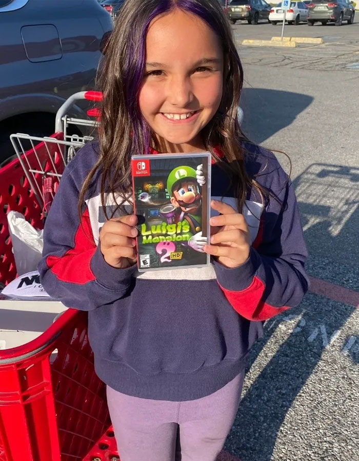 Young girl smiling and holding a Luigi's Mansion 3 Nintendo Switch game outside near a shopping cart and cars.