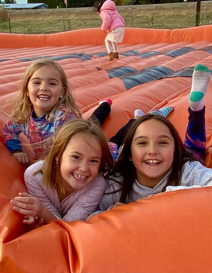 Three smiling girls playing on an orange inflatable bounce house in a grassy outdoor area before being found lifeless.
