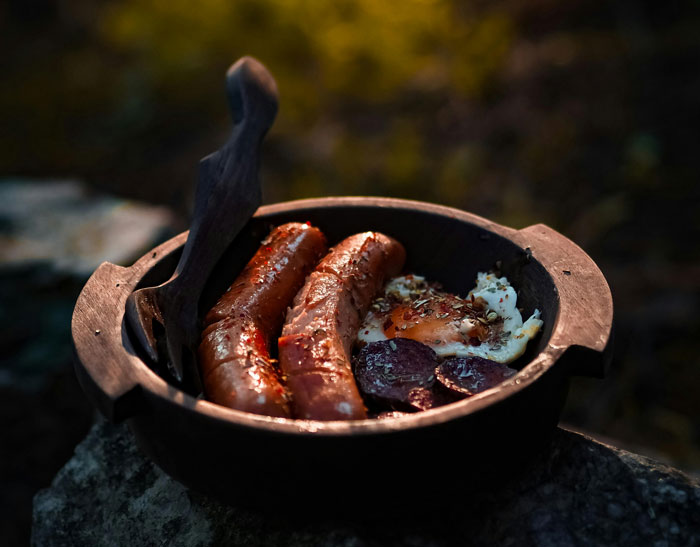 Cast iron skillet with sausages, fried egg, and sliced meat cooked outdoors, illustrating effective old wives' tales cooking methods.