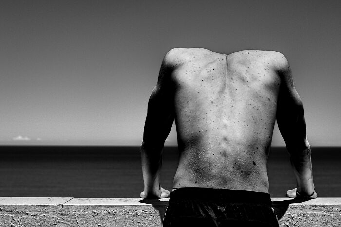 Black and white street photo of a shirtless man leaning on a wall overlooking the ocean, showcasing perfect timing and composition.