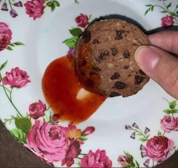 Chocolate chip cookie being dipped into a red sauce on a floral plate, a cursed picture of food that might make you go eeeeuw