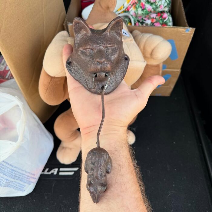 Hand holding a vintage cat-shaped cast iron door knocker, surrounded by stuffed toys and boxes of secondhand finds.