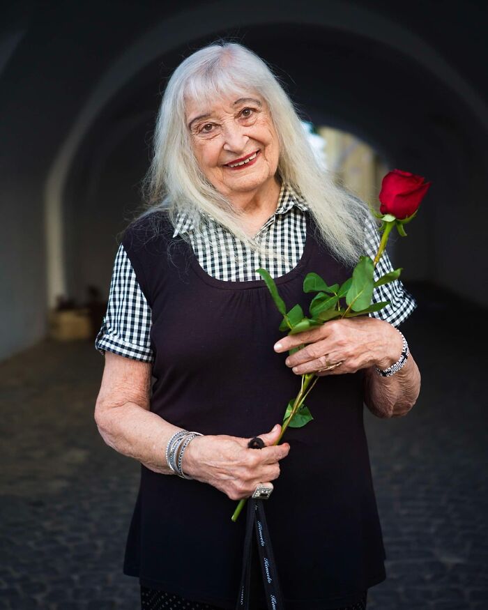 Elderly woman smiling and holding a red rose, showcasing timeless beauty and grace in an outdoor setting.