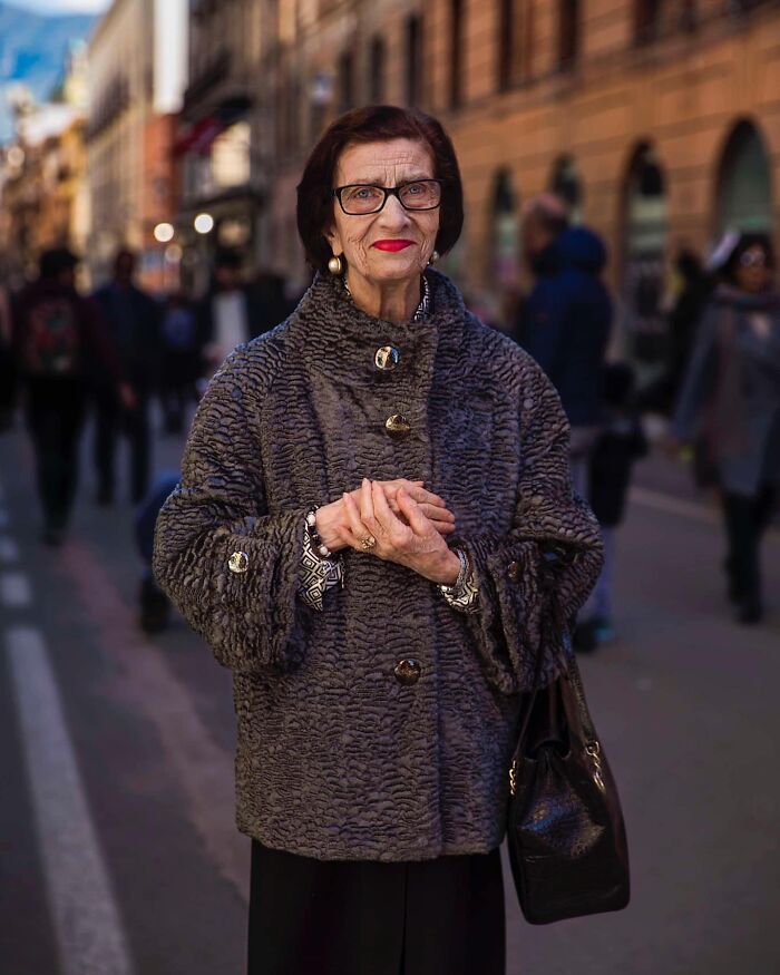 Elderly woman wearing glasses and a textured coat, standing confidently on a busy city street, showcasing timeless beauty.