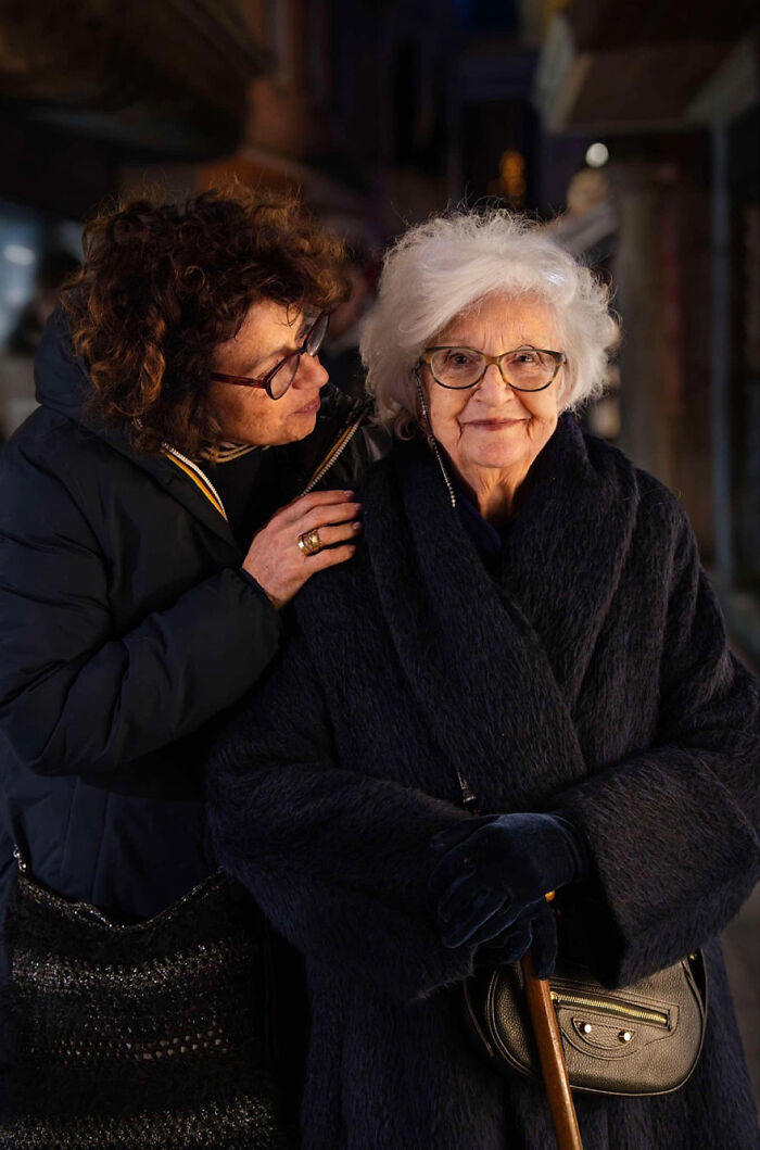Elderly woman with white hair and glasses wearing a dark coat, accompanied by a younger woman in a warm jacket outdoors at night.