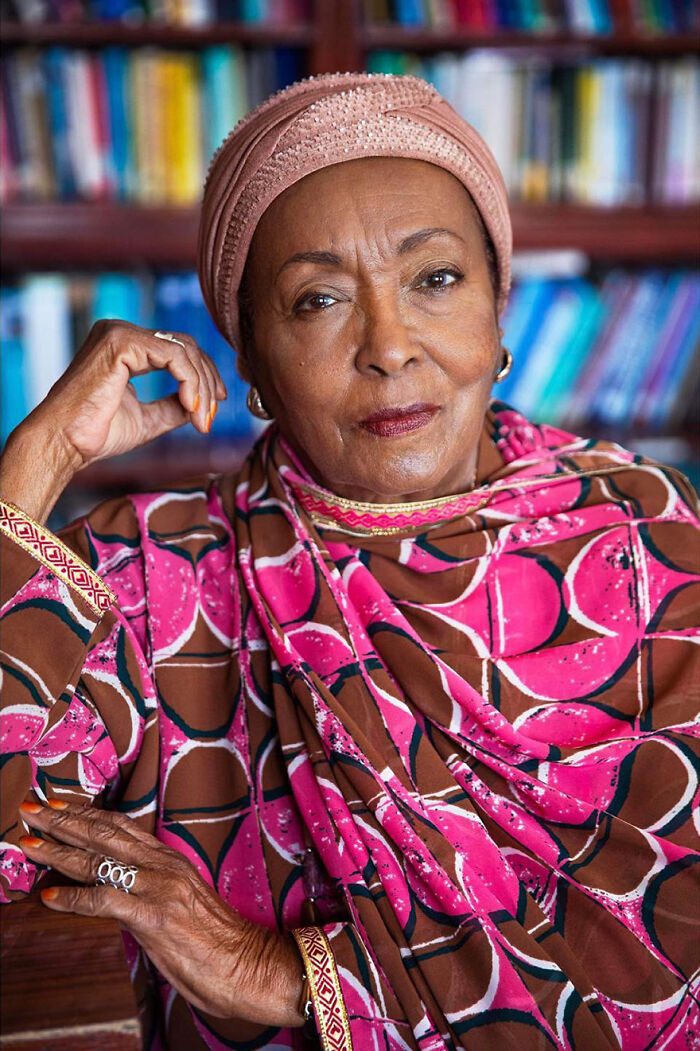 Elderly woman wearing a pink patterned headscarf and dress, sitting confidently in front of a bookshelf indoors.
