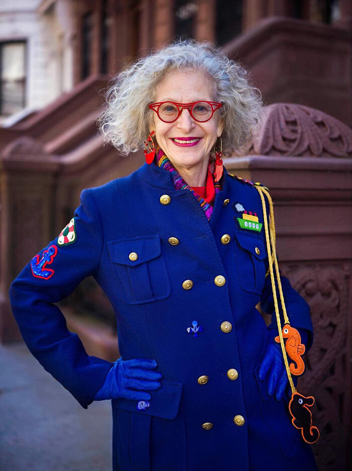 Elderly woman with curly gray hair wearing a vibrant blue coat and red glasses, embodying beauty and style.