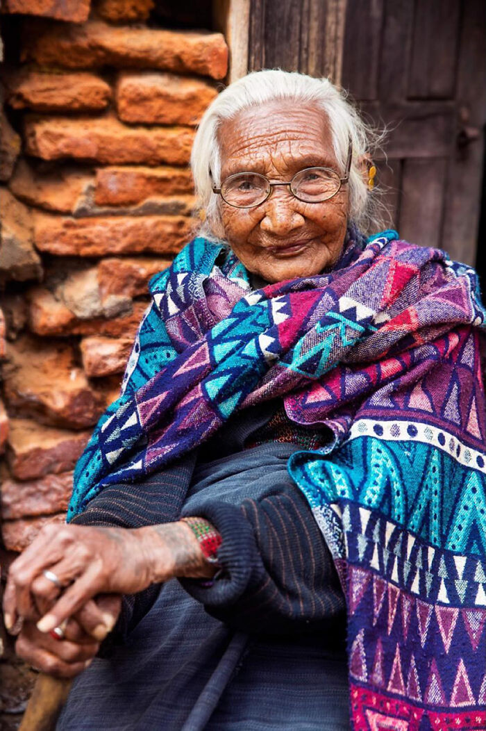 Elderly woman with white hair wearing glasses and colorful patterned shawl sitting against a rustic brick wall.