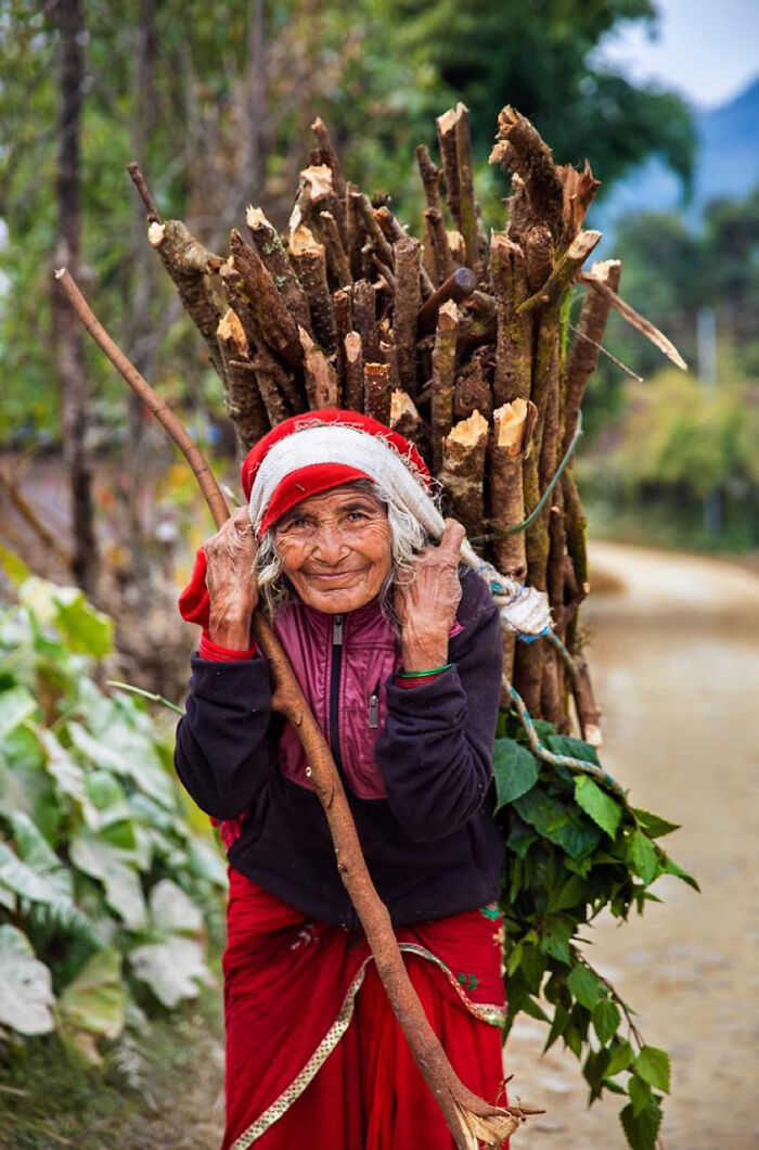 Elderly woman from a rural area carrying a large bundle of firewood, showcasing beauty and strength worldwide.