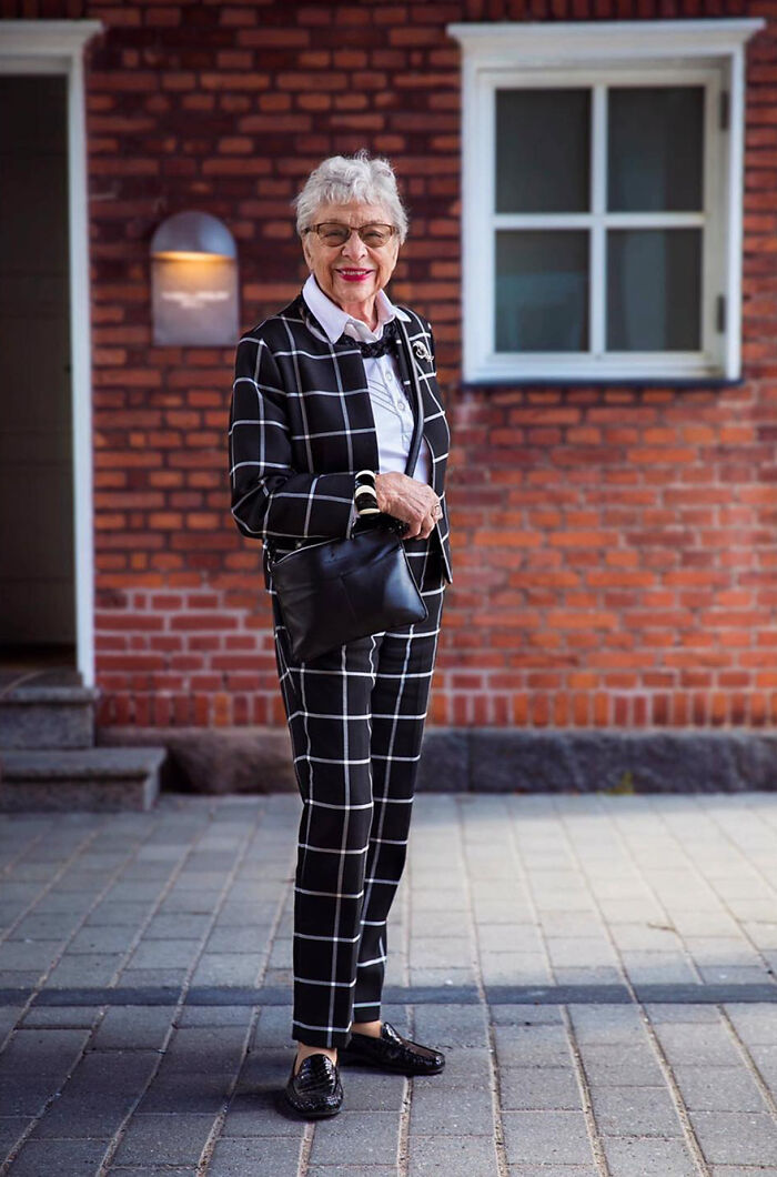 Elderly woman wearing a stylish black and white checkered suit, smiling confidently outdoors against a brick wall.