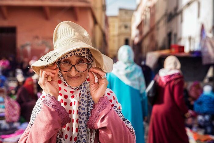 Elderly woman smiling warmly in a sunhat and glasses at a vibrant outdoor market, showcasing beauty and confidence.