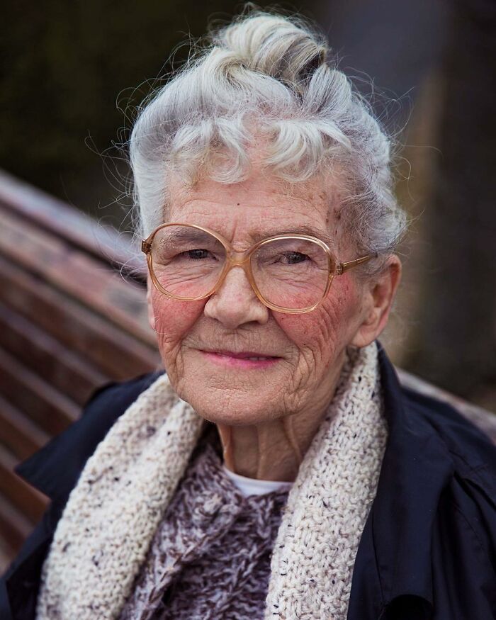 Elderly woman with white hair and glasses smiling gently wearing a knitted scarf and dark jacket outdoors on a bench