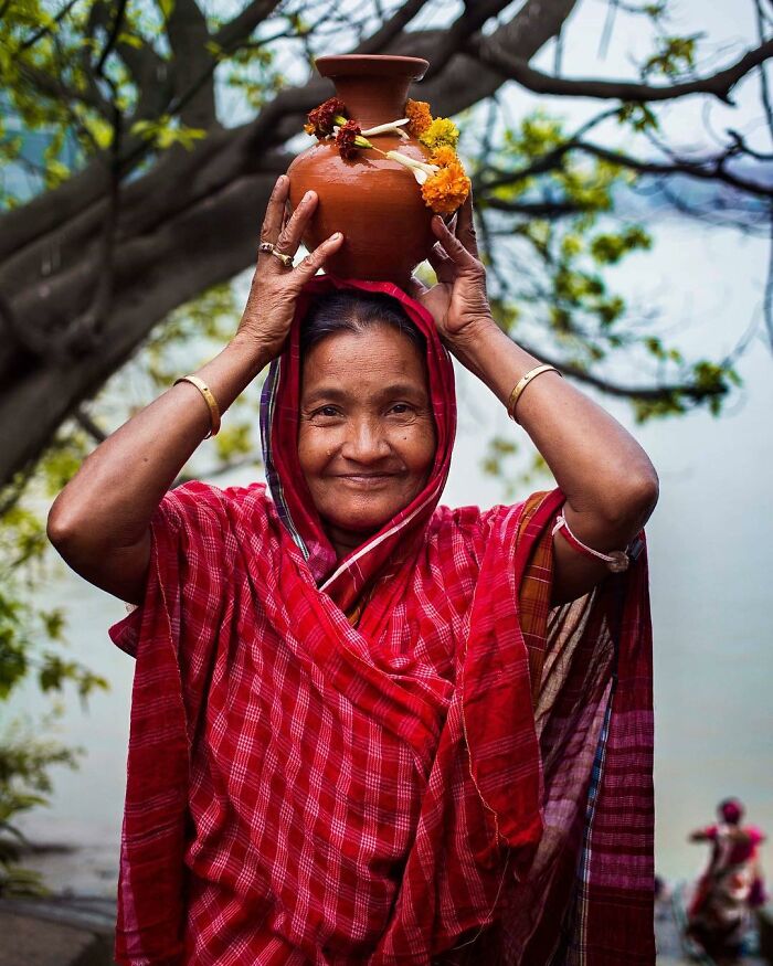 Elderly woman in traditional attire smiling while carrying a decorated clay pot on her head, showcasing beauty and culture.